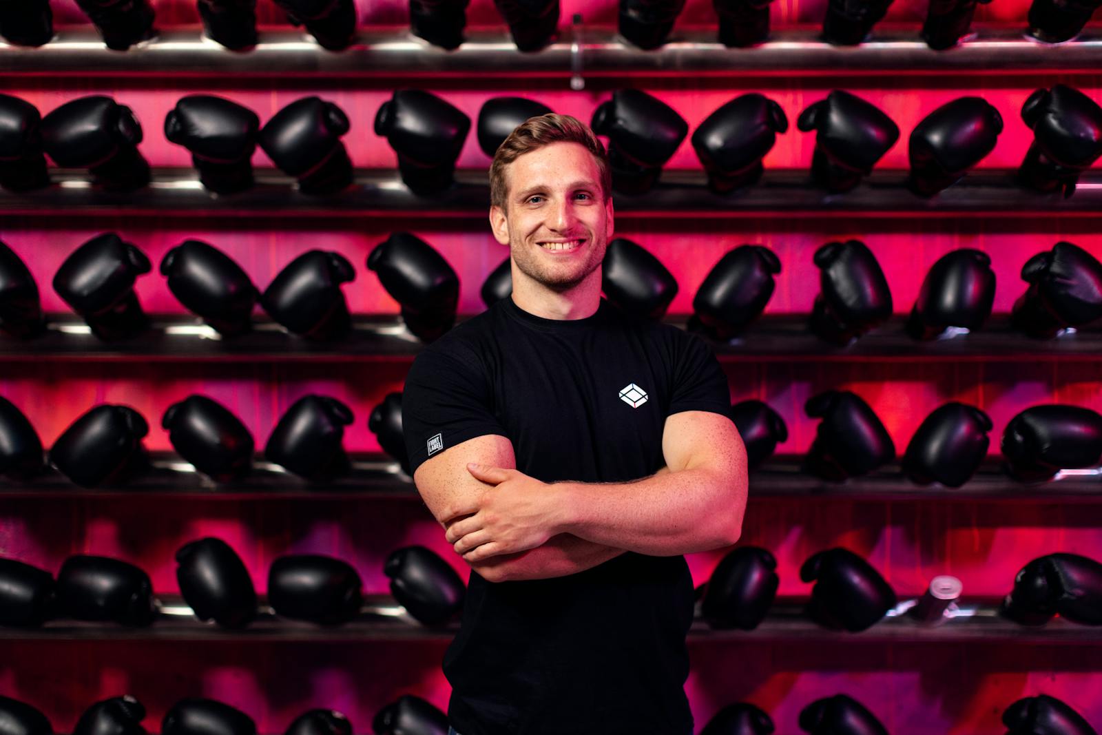 A smiling man in a gym stands confidently in front of a wall of boxing gloves.